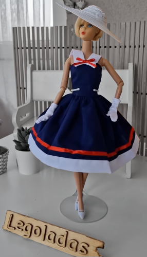 Blonde Hair Fair mod Barbie head on made to move body, posing in a navy blue fit-and-flare dress with red and white trim, including a wide white collar with a red bow and two bands around the skirt bottom, plus white gloves (look like elbow-length but pushed down), wide-brimmed white straw-type hat, white shoes with pointed toes. In the foreground a wooden sign reads "Legoladas."