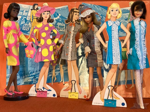 The Francie doll from the "Floating In" reproduction set wears the Sun Spots dress and shoes next to a Francie paper doll in Sun Spots; A vintage Francie doll and paper doll both model Side Kick; and a vintage Casey and Casey paper doll (brunette and blonde, respectively) both wear Iced Blue--though the paper doll is missing her tights and shoes. In the background, the set's folder can be seen. Close inspection of the folder shows illustrations of Francie and Casey in Sun Spots and Side Kick; Francie in Borderline; Casey in Quick Shift; and Francie in the Barbie Fashion Studio Tour. The rest of the folder is obscured by the posed dolls.