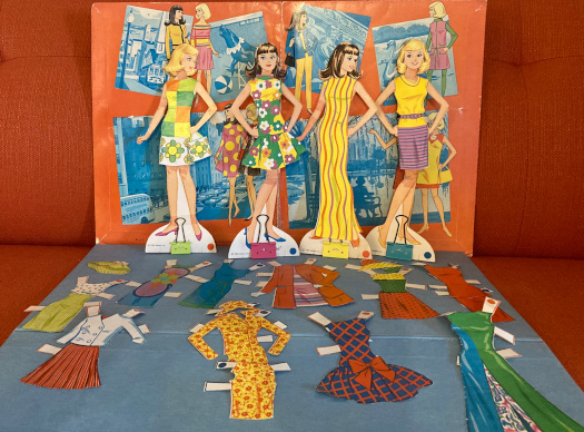 Two Francie and two Casey paper dolls are shown wearing the 1967 Color Magic outfits Pretty Wild, Bloom Bursts, Stripes Away and Smart Switch. Lying around their feet are 1966-67 outfits that came with them, but would normally fit Barbie: Lunch on the Terrace, Sunflower, Junior Designer, Fashion Shiner, Studio Tour, Music Center Matinee, International Fair, Travel Togethers, Beau Time, and Patio Party. The folder propped up behind them also shows, among its illustrations, Casey in Tropicana.