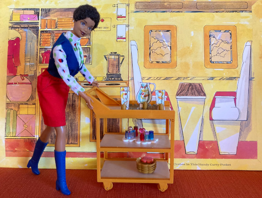 The folder for the 1973 Barbie's Friend Ship is used as a backdrop for a Barbie doll diorama. On the folder, a commercial aircraft interior from a bygone age is illustrated. On the left side is a service station showing a coffee pot on a range, towels, a first aid kit, stacked dishes, and storage for luggage and coats on hangers. On the right side, a booth with small table sits alongside small windows looking out at puffy clouds. A doll in air hostess uniform is posed with a service cart in front of the folder.