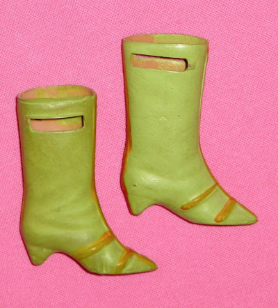 The boots from Barbie's Braniff boarding outfit displayed on a pink background. The boots themselves are a sort of sea green with matte gold accents, both colors painted onto tan colored plastic visible through horizontal slats near the tops of the boots.