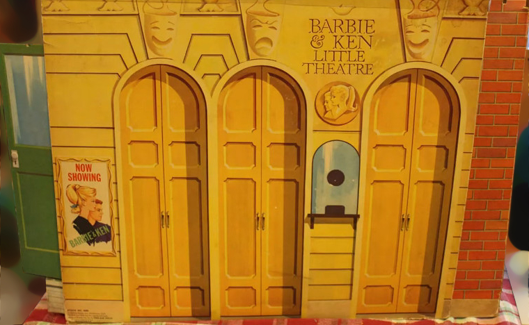 The chipboard box containing the Little Theatre looks like a somewhat cramped building facade, with three sets of double doors in very close proximity, an impossibly small ticket window, and a poster that reads, "NOW SHOWING: BARBIE & KEN." Near the top, the words "BARBIE & KEN LITTLE THEATRE" look like they may be engraved in stone and are surrounded by comedy and tragedy masks. Around the corner to our left we can see a plain green stage door with a glass pane (just illustrated) on the top half.