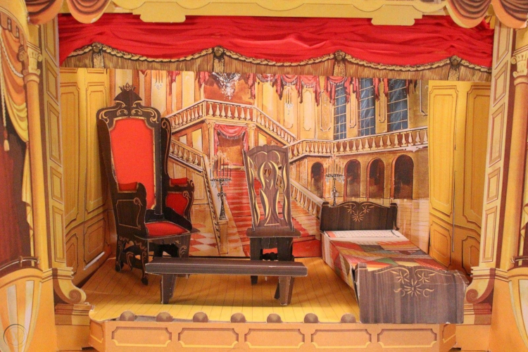 The interior of the Little Theatre playset can be seen. The stage's backdrop consists of a red-carpeted staircase surrounded by a chandelier, candelabras, many arched doorways and soaring windows. On stage, a throne with red upholstery, a simpler wooden chair, a plain wooden bench, and a bed with carved wooden headboard and patchwork quilt are displayed. Along the top, the red cloth curtain is gathered. The wood-look flooring forming the surface of the stage is visible, as is a row of footlights along the front of the stage.