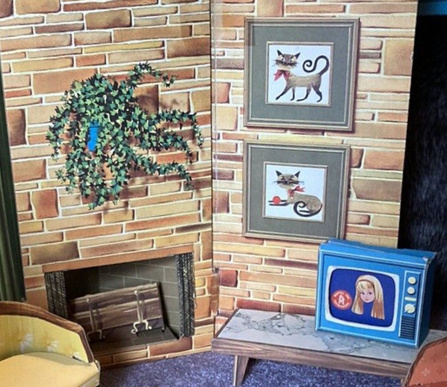 Chipboard interior including fieldstone wall and fireplace (no fire) with potted plant hanging above and two stylized cat drawings hanging alongside. Atop a "marble"-topped coffee table, Skipper is shown on TV. A yellow chipboard armchair peeks in at the edge of the frame.