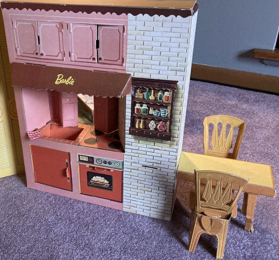 Close up on the kitchen in a current eBay listing. A pass-through window's shutters stand slightly ajar above the range and sink. Above that is a range hood with a Barbie logo, and above this are two small cabinets with four total doors. Below the sink is a cabinet and below the range is an illustrated oven, both of which open and close. A spice rack is illustrated on a column of white-painted brick to the right. A two-legged table extends from the column with two chairs.