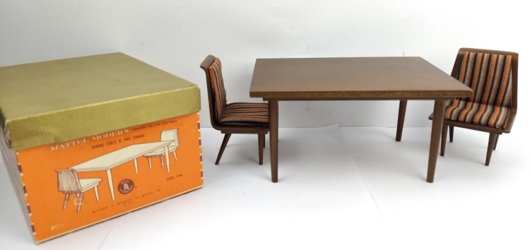 Rectangular wood table with tapered lefts and two chairs. Chairs have no arms, angled back legs, and striped brown/orange/cream upholstery on back and seat. To one side, an orange box with white and brown printing and gold lid has an illustration of the furniture on the side facing the camera, and horizontal stripes on another side that is just in view. The words "Mattel Modern" can be discerned on the facing side.