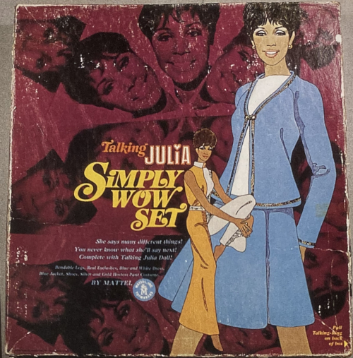 Two Julia characters are illustrated on a square box lid. A smaller figure wears a bellbottomed, belted jumpsuit that is half gold and half silver; a closeup figure wears a teal Chanel-type suit in teal with gold trim with white shirt. In the background on the packaging is a kaleidoscopic collage of Julia's (Diahann Carroll's) photographed face repeated. Text says "Talking JULIA Simply Wow Set" with smaller text explaining that she says many phrases unpredictably.