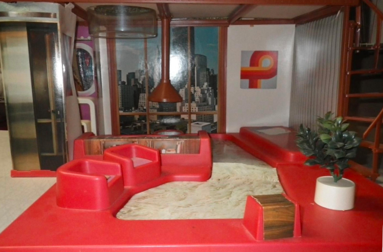 View facing in to the penthouse interior. The inverted skylight can be seen hanging down into the room; beyond, the fireplace hood sits midway across three panels of floor-to-ceiling windows showing a photographed view of the city by day; to the right is a piece of abstract art in typical seventies colorbars/curves. To the left an elevator is represented by a photograph on one exterior wall of a dressing room which is barely visible beyond and includes a "real" oval mirror and nested vanity and tool in white plastic with curved corners. A continuous piece of molded red plastic forms the interior, with two armchairs in the foreground molded facing away from the tall windows. Wood paneling of the electronics console can be seen beyond them, blocking the view of the couch which faces the fireplace and windows (again, all molded in place). In the very foreground is a little stool or table in the same wood paneling, and a potted plant with white plastic base sits to one side (not molded in place, and could also enhance the scene on the rooftop).