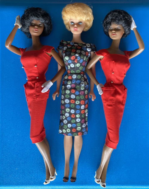 On a bright blue background, two Black Barbies in red sheaths with white gloves and white open-toed heels stand in the "photo finish" pose, with one bent arm to the hip and one to the hair. Between them, a blonde bubble cut with pink lips wears a dark, sheath with multicolored "apple" designs all over and dark open-toed heels. Dramatic uplighting prevents their bubbles from shading their eyes but adds a sense of foreboding to the scene. 
