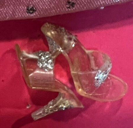Close up on clear plastic Barbie shoes, open-toed with high heels and silver glitter glued across the uppers and all along the heel, still attached to the package which is medium pink in color.