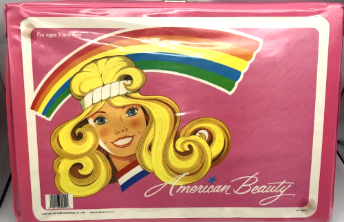 A pink lunchbox features a drawing of the face and hair of a woman with Farrah Fawcett smile and hair, wearing a white sweatband, with red-white-and-blue striped collar. Behind her is a rainbow. Text reads "American Beauty."