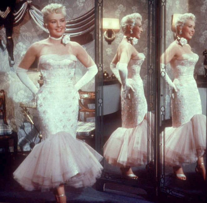 Film still of actress Betty Grable posing before a set of mirrors, wearing an all-white, strapless, beaded mermaid dress, white elbow-length gloves, white open-toed shoes, and white rose tied at the next like a choker. Unlike Solo in the Spotlight, the hemline sweeps up in the front so that the tight skirt ends before her knee in front, changing over to flounce--she can walk in it, in other words.