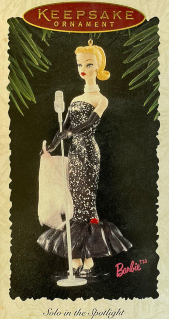 Package exterior for ornament is a photograph of molded and painted Solo in the Spotlight Barbie with blonde ponytail, holding a scarf of real fabric that appears a little frayed even in the product image. Barbie's painted face has striking angles and contrast suggesting the earlier ponytails, but with healthy skin tone. The "glitter" of the dress appears to be painted on in silver.