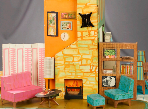 Open floor plan set; at center, a narrow piece of orange wall with a fieldstone fireplace in orange and green stone, with bright fire illustrated inside. Also on this wall are a clock, a wall sconce and some framed art pieces. To either side are a room divider and wood-look shelves with books and TV. Before these are a pink two-seat sofa, coffee table with tall-shaded lamp, teal patterned chair and matching ottoman.