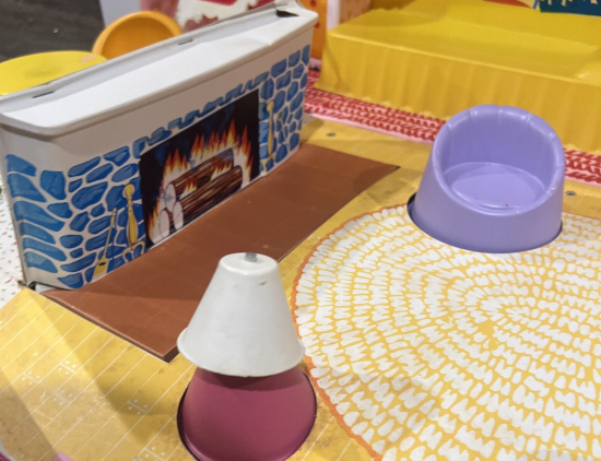 Low plastic fireplace in blue stone, with warm fire illustrated. Before in on an illustrated round, woven rug sits a light purple barrel chair, and table lamp on barrel-shaped table sits to the other side.