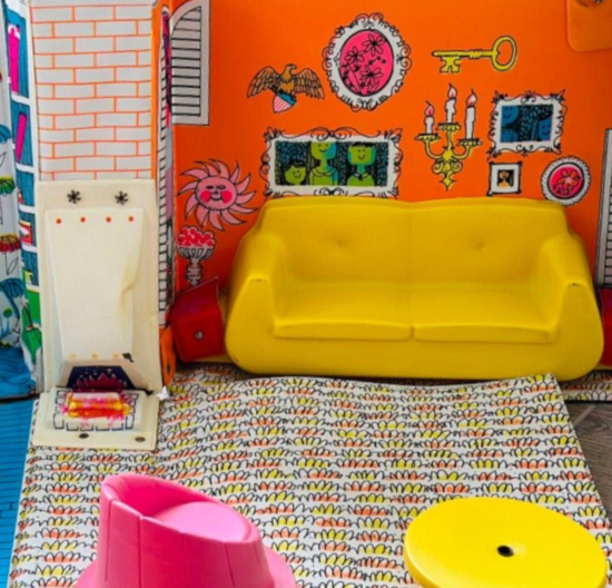 Living room scene in vinyl house. The back wall is fluorescent orange with various illustrated wallhangings in bright colors, including an oversized key, candelabra, eagle, several framed pieces. Beneath these is a yellow plastic couch with two cushions, and before it a vinyl floor piece with deep pile carpet illustrated by rounded scribbles. Left of the orange wall is a narrow section on white brick with, from the height of the couch back down almost to the floor, a nose-shaped protrusion in white plastic. On the underside and beneath are a fire illustrated. In the extreme foreground is a pink barrel chair and yellow-topped table.