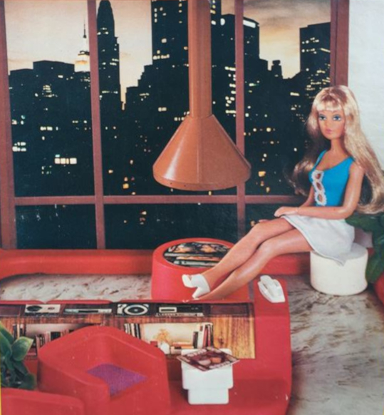 Photograph from product packaging shows Tuesday Taylor doll on a white plastic stool, sitting near a freestanding fireplace in the round, with circular lower half and detached, cone-shaped hood hanging from the ceiling. Behind them, the back wall consists of floor-to-ceiling windows showing city skyline (photograph) at dusk. In the foreground is more molded plastic furniture in red and photographed shag carpet for the floor.