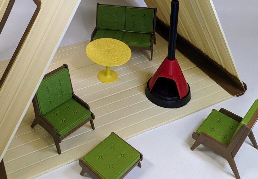 White or off-white plastic A-frame cabin with wood-look accents and open front and back. Wood-look plastic seating with green "cushions", yellow tulip table, and red conical free-standing fireplace sit within and before the house.