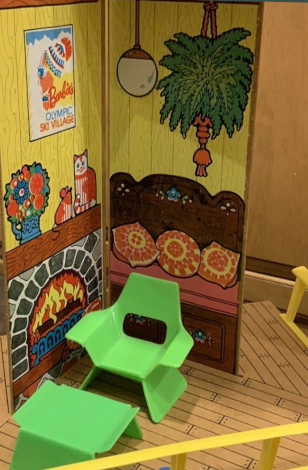 Chipboard two-panel backdrop, green plastic armchair and matching ottoman, on a wood-plank-look floor with yellow railing surrounding. On one panel of the backdrop are illustrated a wooden bench with cushions and alpine flower detailing, hanging house plant and pendant lamp. On the other are grey stone fireplace with bright fire, and on the wooden mantel a jar of flowers, cat and mouse (statuettes? animals that are friends?) all beneath a "Barbie Olympic Ski Village" poster. The resat of the wall is yellow with wood-like lithographed texture.