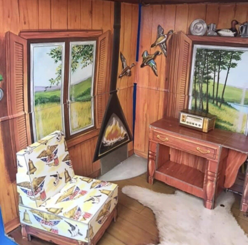 Chipboard house interior. On two walls are windows illustrating rural scene, and between them, a conical fireplace in freestanding style, with roaring fire inside. Nearby are a patterned chipboard chair and desk or sideboard with radio atop. The walls and floor are wood-panel look, with a bearskin rug illustrated on the floor.