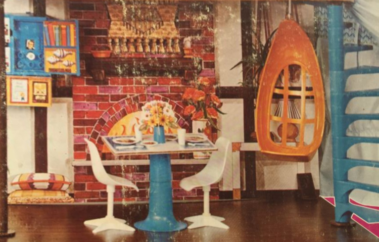 House interior with red brick wall section including semicircular opening and blazing fire within. In front of this, a square table and two chairs in while and blue plastic. To one side hangs an egg-shaped orange plastic chair and a blue spiral staircase leads to the upper level (not shown).