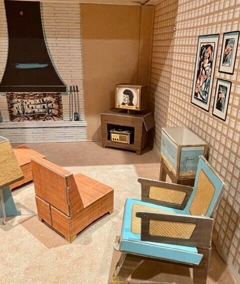 Chipboard living room. On the back wall, a fireplace in white brick with black hood dominates. Next to it, a TV stand with black-and-white TV showing a smiling woman's head; she looks like a dark-haired Betty Grable. Beneath the TV in the stand is a record player. Closer to the camera, a "fish tank" (printed onto chipboard), wood-look armchair, and upholstered-look orange chair with matching ottoman are arranged. One wall has plaid wallpaper and a few indistinct framed pieces illustrated.