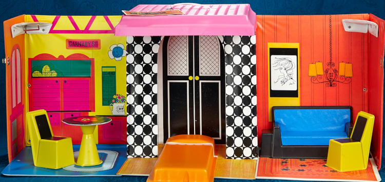 Color photograph of vinyl house interior consisting of 3 rooms. Walls are orange and yellow with some decor drawn on as described in the post. Floors in 3 segments are: blue with a white oval rug, orange with blue and red flowers, and in the center segment a hardwood pattern before a pair of arched black-and-white doors resembling an entryway (like the one in the previous image) but could stand in for a closet. Furniture is an orange molded bed, a black plastic sofa with blue vinyl "upholstery", two yellow plastic chairs with black seat and back, round yellow circular table with black top and red flower design on top. The walls between "rooms" are shallow and printed like black and white tile.
