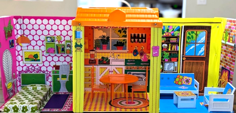 Color photo of vinyl house interior. Same layout of 3 rooms but with full walls between. Left is a bedroom with pink-and-white walls, illustrated with pendant lamp, potted plant, white wicker chairs, green desk or vanity, floating shelves with piggy bank, alarm clock, and other knick-knacks, and framed landscape picture. Green headboard is lithographed on the wall, while the bed itself--white with green flower pattern, is a physical object. Center room is a kitchen with orange walls and, illustrated thereon, white and yellow wood cabinets, dark green oven, hanging potholders and spice rack, and the floor is tiled with circular rug. Orange round table and two high-back chairs sit in the space. Room at right is living room with blue floor, brick fireplace on one wall, wooden door with glass pane (illustrated) and bookshelves with gumball machine, TV, other knick-knacks on second wall, and white plastic sofa, armchair and low table in the room. Sofa and chair have blue "upholstery" stickers and table has mosaic tile-look top in orange, yellow and pink. The house has a flat orange plastic roof. At the front of the walls between rooms is a small section of yellow exterior wall; on one wise is a pink mailbox with "Barbie" in a familiar logo, and the other has a hexagonal-shaped porchlight.