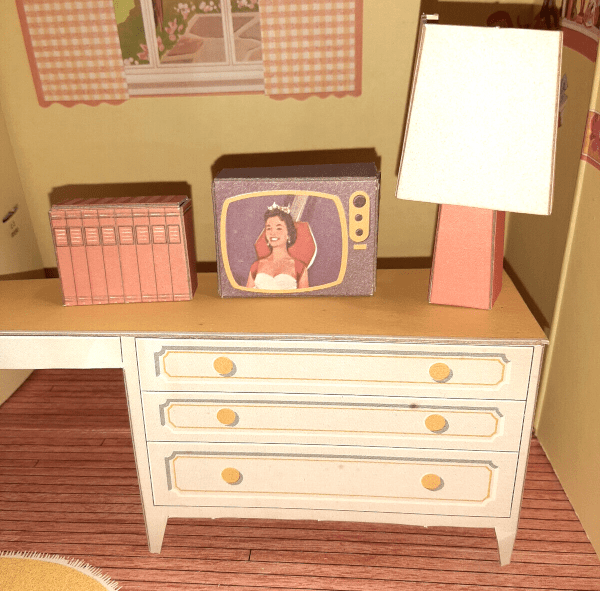 Purplish chipboard TV. On "screen" a dark-haired woman smiles brightly. She wears a tiara, a white gown with sweetheart neckline, and Elizabethan collar in red rising behind her head. She's shown to just below the bust, so we only assume that her garment is a gown; in a pageant scenario it could easily be a bathing suit.