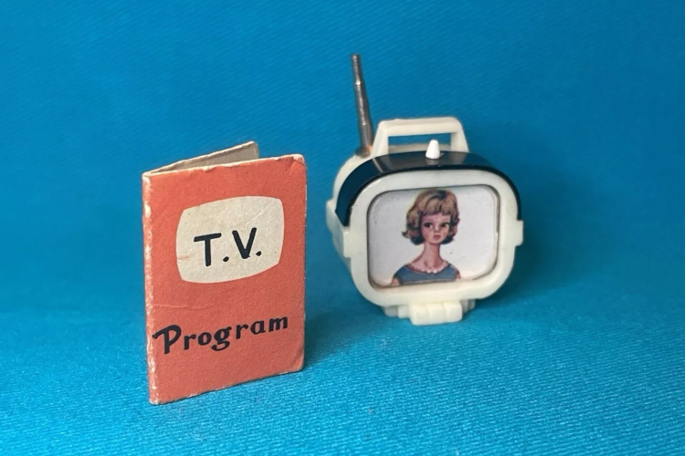 On a blue background, a folded piece of paper or cardboard says "T.V. Program" on a tV-screen-shaped blob with salmon background. Beside the TV program, a slightly ovoid plastic set in white plastic, with a handle and metal-look antenna on top, shows a girl or woman with a dirty blonde shot flip hairstyle and fringe, pearls, and a blue sleeveless top or dress.