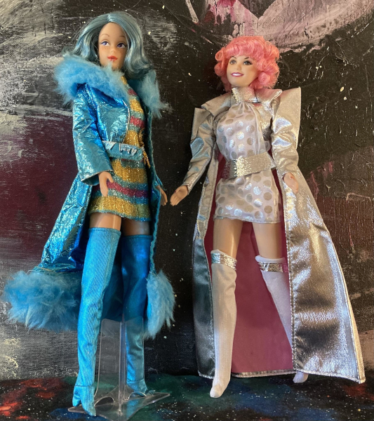 Two dolls pose before an abstract starry-looking background. Left is One Modern Circle Melody, with teal hair, wearing a short, metallic dress of gold, magenta and teal stripes, thigh-high metallic teal boots, and a shiny teal metallic knee-length coat with same-color metallic belt and oversized (for Barbie) "fur" at the collar and hem. Beside her is the pink-haired Frenchy doll wearing a silver-on-white polka-dotted minidress with thick silver belt and neck trim, white fabric boots with silver trim covering the knees, and floorlength silver coat with pink lining and sharp pointed shoulders.