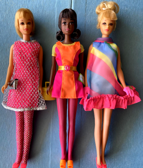 3 Francie dolls lined up on a light blue background. Left is Hair Happenins Francie wearing a pink shift with silver net overdress, pinkish silver net tights, silver clutch, and pink squishy bow shoes; center is Black Francie repro wearing a pink and orange minidress with yellow belt, pink daisy accent at buckle, yellow net undershirt, sheer pink hose, hard yellow purse, and orange platform shoes; right is Growin Pretty Francie in white shift with sheer, rainbow-striped overdress, short pink gloves, and pink Mary Janes or peep-toe shoes.