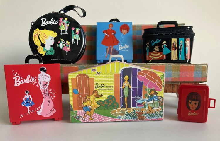 Six miniature Barbie accessories lined up on and in front of the Dream House sofa. At rear, on the sofa, left to right are a piece of black vinyl luggage in a hatbox shape with colorful Barbie illustrations; a blue carrying case illustrated with Titian bubble cut Barbie in Red Flare, a black vinyl boxy shaped luggage piece with ponytail Barbies illustrated in front of skewed rectangles. Front row, left to right: the Enchanted Evening case from the previous image; the mini Barbie Family Deluxe house exterior, with faithful representation of the full-sized house's front cover; and a small red plastic case with American Girl Barbie's head screened on along with the Barbie signature in white.