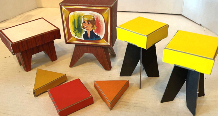Close up of a color "TV" showing a smiling blonde. Plus a wood-look table with white top, two square stools with yellow tops and black bases, and three boxy little "pillows," a square and two triangles in red, orange and mustard color.