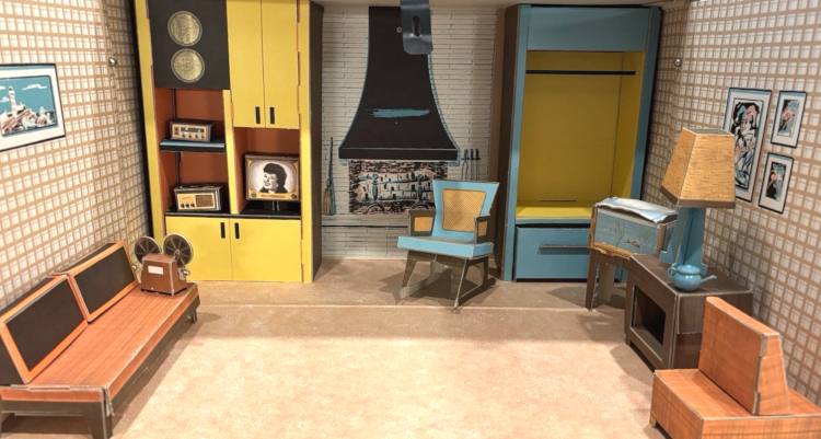 A large (for Barbie or Tammy) room interior. Furnishings are folded chipboard. Furnishings are described in this post. The wallpaper is a brown/orange check pattern on white; the sofa and chair are orange with brown and "wood" accents. Blue and yellow storage units occupy the back corners, one with a rod for hanging clothing and one with various shelves and cabinets, some occupied by the "electronics." in front of the fireplace, at center rear, sits a blue and "wicker" rocking chair. Some indistinct paintings hang on the walls, and the floor is wall-to-wall carpet texture in beige.