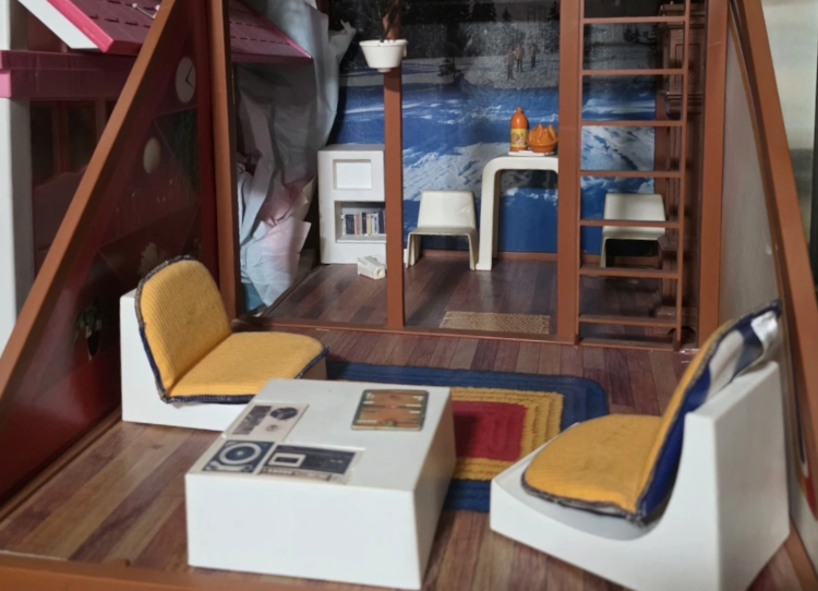 Photograph of the lower level of the ski chalet version of the house. Furnishings are of white plastic with colorful cushions, a rainbowy area rug probably printed on the wood-look floor, and a plastic plant in a hanging white pot.