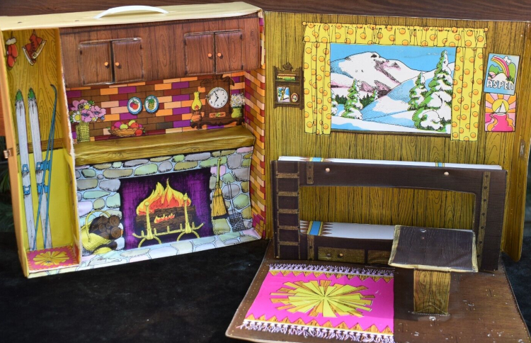 Photo of interior of the Mountain Ski Cabin, essentially as described. On the mantel alongside the clock (these are all illustrations on the vinyl wall) are a vase of flowers, a bowl of fruit, and a couple little pictures of fruit. Along the top are little cabinet doors in the vinyl that really open and close. To the left of the fireplace, spanning from floor to ceiling, is an inset closet with skis, skates and mittens illustrated. On the wall to our right, to either side of the window are a little wooden organizer of something, and a couple posters, one saying ASPEN with a rainbow and one showing a sailboat in the sunset. The rest of the room folds down from this wall, consisting of the bunked with small ladder, the table that extends from them, and a wood-look floor with a pink and yellow throw rug.