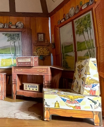 Closeup on chipboard furniture inside the folding one-room house. The chair has a screening including the US Revolutionary War symbolism listed in the main text--the eagle is golven and clutches a stars & stripes bunting--plus a side table with audio equipment similar to that included in the previous structure, only wood-look instead of bamboo. On two visible walls are "windows" onto a lake, and shelves overtop of the windows are lined with silver plates and tankards and multiple decoy ducks. Screened on the floor is a white bearskin rug.