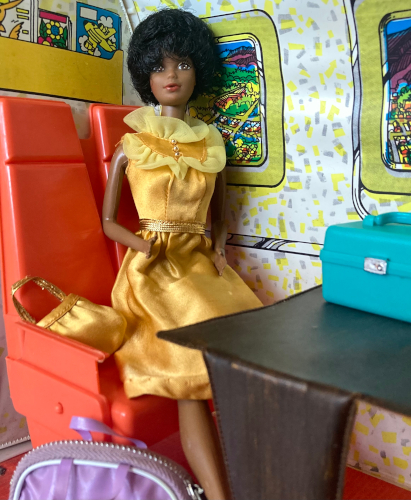 1980's first Black Barbie sits in a row of two orange plastic seats with a brown wood-look table before her. Along the wall are small rectangular windows showing illustrations of the ground below an airborne plane. Barbie wears a gold sleeveless dress that just covers her knees, with a metallic gold belt, ruffles and a row of buttons at the collar, and a matching purse with metallic gold handle. On the table before her sits a teal train case with silver hardware. On the ground next to her feet is a light purple zippered bag with two handles, partially cut off at the bottom of the shot.