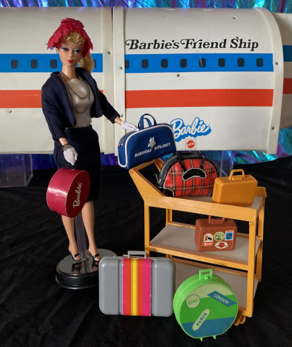 Blonde Commuter Set reproduction Barbie stands holding her hat box in one hand, the other hand resting on Barbie's American Airlines flight bag, which in turn sits on the edge of the Friend Ship cart. The exterior of the Friend Ship looms in the background, with the appearance of a row of porthole windows. Also lined up on the cart are the Winter Holiday plaid overnight bag, a small gold-colored case that came with the Friend Ship, a second small, darker brown case covered in travel stickers that came with Busy Barbie and friends. In front of the cart sits the aluminum-look suitcase from the Star'Vette and a round green hat box.