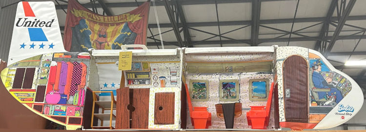 A second view of the opened Friend Ship withough dolls. The cart is parken in the open space next to the galley, while the empty hanger rack visible above it. Two rows of two orange plastic seats sit facing each other over the table with three windows beyond. Ona vinyl panel that swings open at the front, a captain character is illustrated sitting in the cockpit. Behind him an arched dark wood-look door in the vinyl can open and close but doesn't lead into another space for play, just out the back of the vinyl panel.