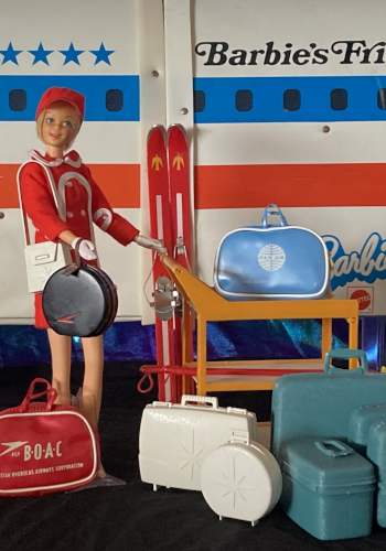Photograph of blonde Casey doll wearing a red Shillman minidress with white trim and red het. She carries luggage, pushes a cart loaded with more luggage, and is surrounded by luggage. At back a pair of skis rest against a red, white and blue structure that is mostly not in the shot, but some letters are visible spelling "Barbie's Fri..." over a row of porthole windows.