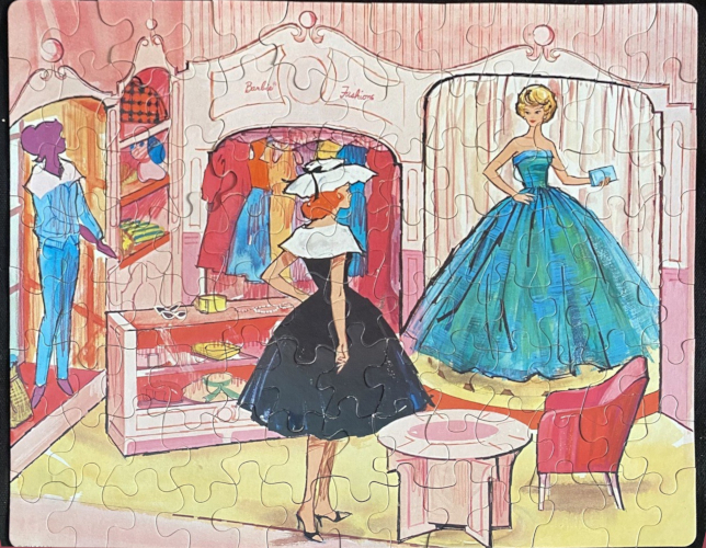 Another assembled 100-piece puzzle, this one shows Barbie's Fashion Shop. On the left side is a window with a mannequin wearing a blue jacket and slacks. Next to that are some shelves holding bags, a stuffed dog, and other accessories; a glass case holding hats with two pairs of white sunglesses on top; and a clothing rack filled with colorful dresses and maybe a red flare coat. Midge stands in the foreground with red hair, wearing a black fit-and-flare dress with wide white collar, a wide-brimmed white hat with black ribbon, and black open-toed shoes. Barbie, another blonde bubble, stands on a low stage before a white curtain, wearing a full-skirted ballgown in blue and green and holding a powder blue clutch. The scene is completed by a circular table and red armchair. There is a pleasant softness to the drawing style, while overall it's a highly accurate depiction of the real Fashion Shop.