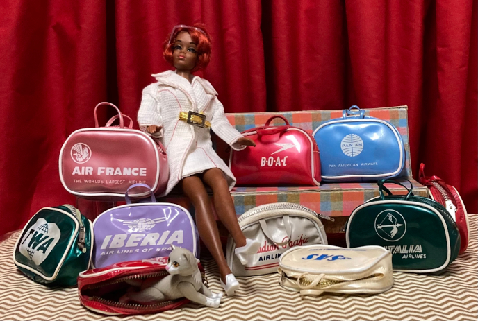 Christie with oxidized hair wears the white jacket, gold belt and white pilgim shoes of White 'n' With It. She sits at one end of the plaid sofa and is surrounded by soft-walled vinyl bags with two handles, zippers, white piped trim, and airline logos, most printed in white. They are for: Air France, in a frosty pink; B.O.A.C., in glossy red; Pan Am, in sky blue; AZlitalia, in dark green; SAS (Scandinavian Air) with blue test on a sheen-y white; Airr Iberia on lavender; and TWA on the same green as Alitalia. An additional red bag we can't read sits sideways off the end of the couch, and one more unreadable red bag lies open on its side, unzipped. Kiki is halfway out of this bag, arms stretched to the right but head turned gazing to our left. She looks spirited. Just between us, One of the red bags is a second BOAC and the oher was one of the less-glamorous domestic airlines, so I didn't try to make them clear in the shot. 