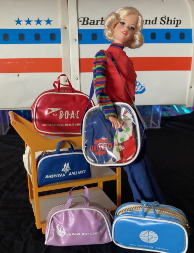 With the Barbie's Friend Ship Plane as a backdrop and black "tarmac" beneath, blonde short flip Stacey reproduction stands next to the Friend Ship's service cart, wearing the United Airlines flight attendant uniform's blue slacks and red shirt with long striped sleeves. Over her shoulder is a largish square bag with a clear fron on which the United logo can only faintly be read because of the colorful contents within: navy skirt, white shirt with "wonder bread" polka dots, and red vest. On the service cart, top shelf is the second red BOAC bag. While the one isn the previous image just said BOAC in large letters with a swoosh logo, this one says "fly BOAC" with the swoosh, then spells out British Overseas Airline Company below. Middle shelf holds the Barbie American Airlines bag, which is canvas and not vinyl like the other bags. On the ground in front of the cart is a lavender bag for Japan Air Lines and the sky blue Pan Am bag.
