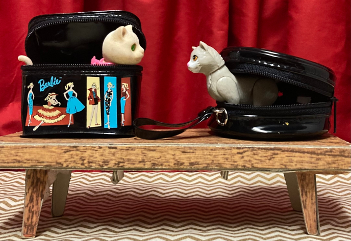 The two Hallmark cases sit unzipped on the wood-look coffee table from Barbie's first Dream house. On the right the hat box lies on its side, partway open, Kiki peeking out. To the left the train case lid is open and a white cat with green eyes and pink bow looks out (a 90s Barbie cat). The two cats have locked eyes from their separate cases. It doesn't necessarily appear friendly. 