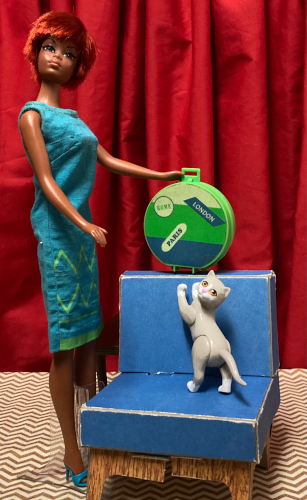 Oxidized Julia wears the Junior Designer turquoise sheath with turquoise open-toed heels. She stands next to the blue chair. In her hand and supported by the back of the chair is a green plastic hat box with white, green and blue stripes and sticker-like details that say "Rome," "London," and "Paris." Kiki stands on the chair, her front paws against the back of the chair, like she's reaching toward the hat box. Both Kiki and Julia have their heads turned back toward the camera as if they've been interrupted.