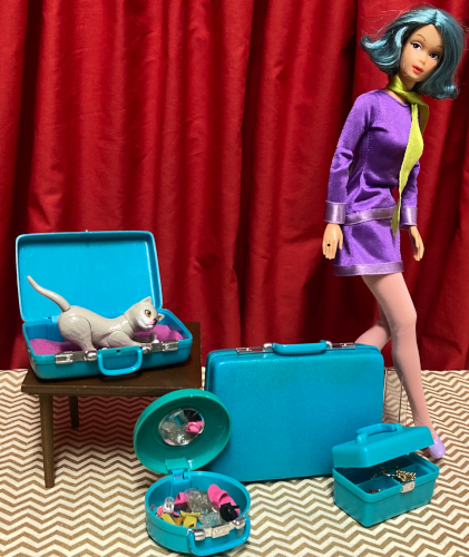 One Modern Circle Melody, with mod Barbie's face mold, teal short flip hair, and a purple mini dress and green scarf from Scooby Doo's Daphne, looks like she's walking through the frame. Teal plastic luggage sits nearby, some open, some standing upright. These pieces are a brighter hue than the earlier Samsonite Silhouettes, and they have the appearance of metal closures and handle attachments. On the floor, a train case sits slightly open with jewelry visible in a compartmented top tray. A hat box sits wide open, a circular mirror visible in its lid, full of open-toed shoes of carious hues (I panicked and couldn't think of any good hats for this scene). A large suitcase sits upright, closed. On the small brown suitcase a slightly smaller suitcase sits open, a bit of magenta material within, and Kiki also within, kneading the material.
