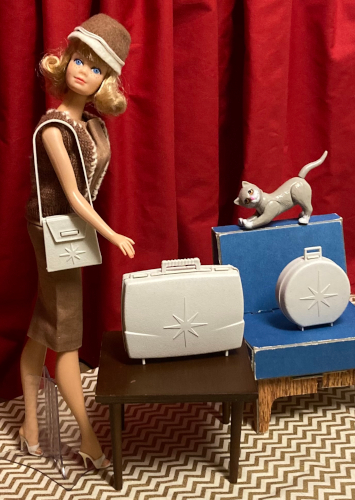 Midge wears Sorority Meeting: a brown sheath, matching knit brown vest with off-white trim, white open-toed heels, and brown felt hat with off-white trim. Over her shoulder hangs a hard plastic off-white purse adorned with a molded sunburst. In front of her is the wood table holding an off-white hard plastic suitcase, also with a sunburst emblazoned on the front. Beside the table is the blue chair from Barbie's Dream House. A hard plastic off-white hat box with sunburst sits upright on the chair. Kiki stretches atop the chair back.