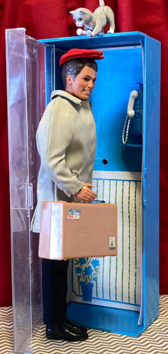 Back at the phone booth. A nineties Ken with brown hair wears Rally day: a red cap and tan jacket, plus a pair of black slacks and black shoes. He stands in the open doorway of the phone booth, turned halfway toward the insde. In his near hand is a boxy tan suitcase with two little stickers, one at the top left corner and one at the bottom right, that are a little colorful but inscrutable. Faintly, the word "Samsonite" is stamped into the center of the case. Atop the booth sits Kiki, posed as if ready to pounce or at least swat Ken's head as he passes beneath.